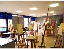 Aula di arte con numerosi cavalletti in legno e tele disposte tra tavoli e sgabelli su pavimento in graniglia. Grandi finestre laterali con tendine blu e illuminazione a pannelli sul soffitto.