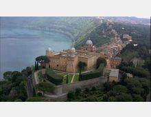 Veduta aerea di un grande complesso storico fortificato con cupole e giardini geometrici terrazzati, circondato da mura e alberi. Sullo sfondo si vedono un lago lungo la costa e un borgo collinare con edifici addossati al pendio.