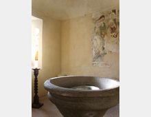 Interno di una piccola cappella con pareti in intonaco chiaro, un grande fonte battesimale in pietra al centro e un affresco religioso parzialmente rovinato sulla parete. A sinistra si vede una finestra con luce naturale e un portacandela alto vicino al pavimento in piastrelle.