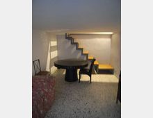 Piccolo interno seminterrato con soffitto basso, pareti bianche e pavimento in graniglia. Una scala a gradini in legno lungo la parete conduce a un soppalco, con tavolo rotondo scuro e sedie al centro e un letto singolo sul lato sinistro.