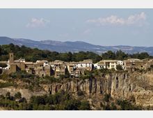 Panorama di un borgo storico con case in pietra e una torre campanaria, disposto lungo il bordo di una scogliera rocciosa. Sullo sfondo si vedono colline e montagne con boschi sotto un cielo parzialmente nuvoloso.