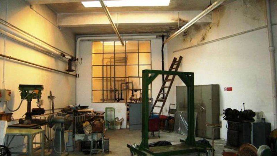 Interno di un’officina industriale con pavimento in cemento, macchinari e banchi da lavoro, tubazioni a vista e lucernario sul soffitto. Sul fondo una grande finestra a griglia e una scala appoggiata vicino ad armadi metallici e attrezzature.