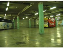 Ampio parcheggio sotterraneo o deposito per autobus con pavimento in cemento lucido, colonne cilindriche verde chiaro e illuminazione a neon sul soffitto. Alcuni autobus sono parcheggiati ai lati, con numeri di corsia visibili sulle travi.