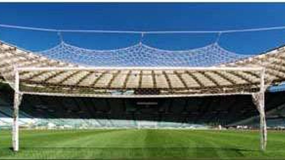 Vista panoramica di uno stadio all’aperto con ampio campo da calcio in erba e gradinate circolari. La copertura curva è sostenuta da pilastri e da una struttura a rete di cavi, con cielo sereno sullo sfondo.