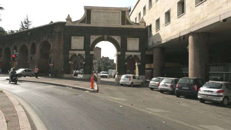 Arco monumentale in muratura con grande apertura centrale e archi laterali, affacciato su una strada urbana con auto parcheggiate e corsie di traffico. A destra si vede un edificio moderno sopraelevato su pilastri, che incornicia il passaggio vicino all’arco.