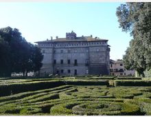 Veduta di un grande palazzo storico in pietra con tetto in coppi e finestre regolari, sullo sfondo di un cielo sereno. In primo piano si estende un giardino formale con siepi basse potate a labirinto, circondato da alberi ad alto fusto.