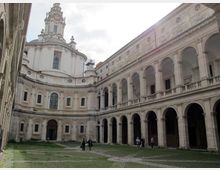 Cortile interno di un complesso storico in pietra, con lunghe gallerie porticate ad archi su due livelli e una facciata curva con grandi finestre. Sullo sfondo si vede una cupola con lanterna e decorazioni barocche, con prato e pavimentazione in pietra al centro.