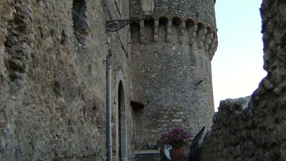 Vicolo stretto in pietra con pavimentazione a ciottoli e muri grezzi, illuminato da una lanterna a parete. In fondo si vede una grande torre cilindrica in muratura con beccatelli e piccole aperture, con un vaso di fiori vicino al passaggio.