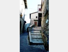 Vicolo stretto di un borgo storico con scalinata in ciottoli e muretto curvo, affiancata da edifici in pietra e intonaco con persiane in legno. Lampioni a parete e una piccola rampa sopraelevata conducono verso una casa con porta in legno.