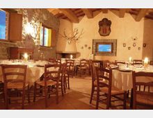 Sala da pranzo in stile rustico con tavoli rotondi apparecchiati e sedie in legno, pavimento in cotto e soffitto con travi a vista. Pareti in pietra e intonaco con camino ad angolo, finestre con scuri in legno e decorazioni murali come trofei e stemmi.