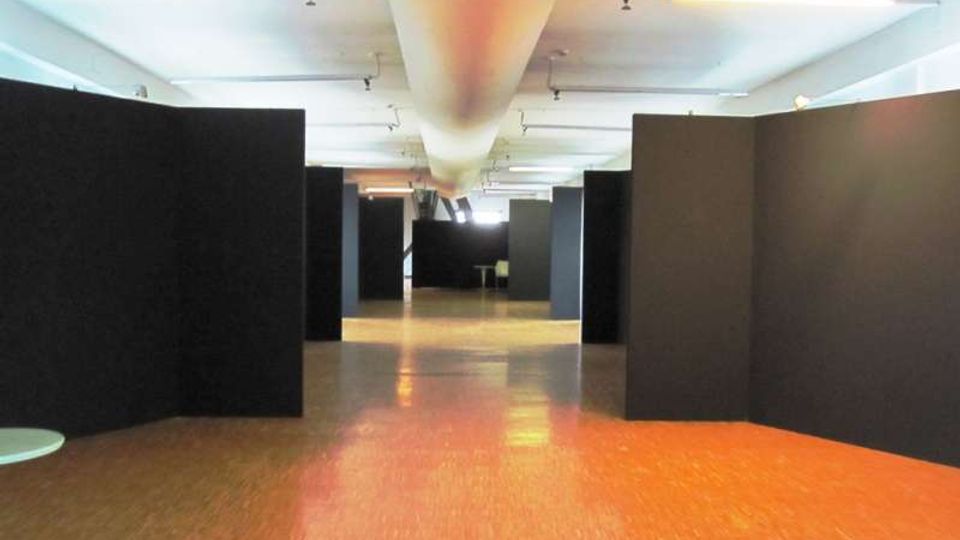 Ampio spazio interno tipo galleria espositiva con pavimento lucido e soffitto basso illuminato da luci lineari. Lungo il corridoio centrale sono presenti grandi pannelli divisori neri che formano aree separate.