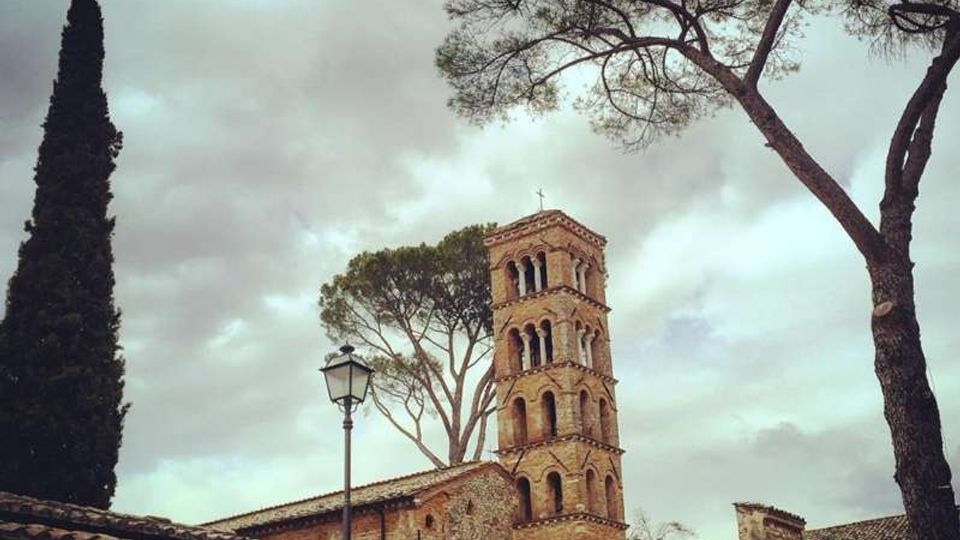 Complesso storico in muratura con edificio basso intonacato giallo e tetti in coppi, affiancato da un campanile romanico in mattoni con archi sovrapposti. In primo piano si vede un muro in pietra e un lampione, con alti pini e un cipresso sullo sfondo sotto un cielo nuvoloso.
