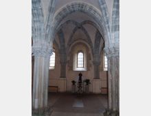 Interno di una chiesa in pietra con archi a sesto acuto e volte costolonate sostenute da colonne. Sul fondo si vedono alte finestre ad arco che illuminano una piccola area centrale con piedistalli e piante.