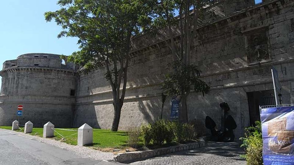 Mura di una fortezza in pietra con bastione cilindrico, affiancate da una strada asfaltata e da un’area verde con alberi e dissuasori in cemento. Accanto a un ingresso ad arco si vedono un’aiuola, una scultura scura e un cartello informativo.