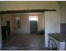 Interno rustico e spoglio con pavimento in cotto, pareti intonacate e una grossa trave in legno a vista che attraversa la stanza. Sul fondo ci sono una porta in legno rialzata da alcuni gradini e una finestra, con un piano in muratura sul lato destro e una sezione di piastrelle decorative.