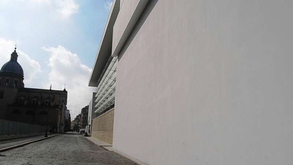 Strada urbana in acciottolato affiancata da un grande edificio moderno con lunga facciata bianca liscia e fascia di finestre orizzontali. Sul lato opposto si vede un edificio storico in ombra con cupola, con cielo parzialmente nuvoloso sullo sfondo.