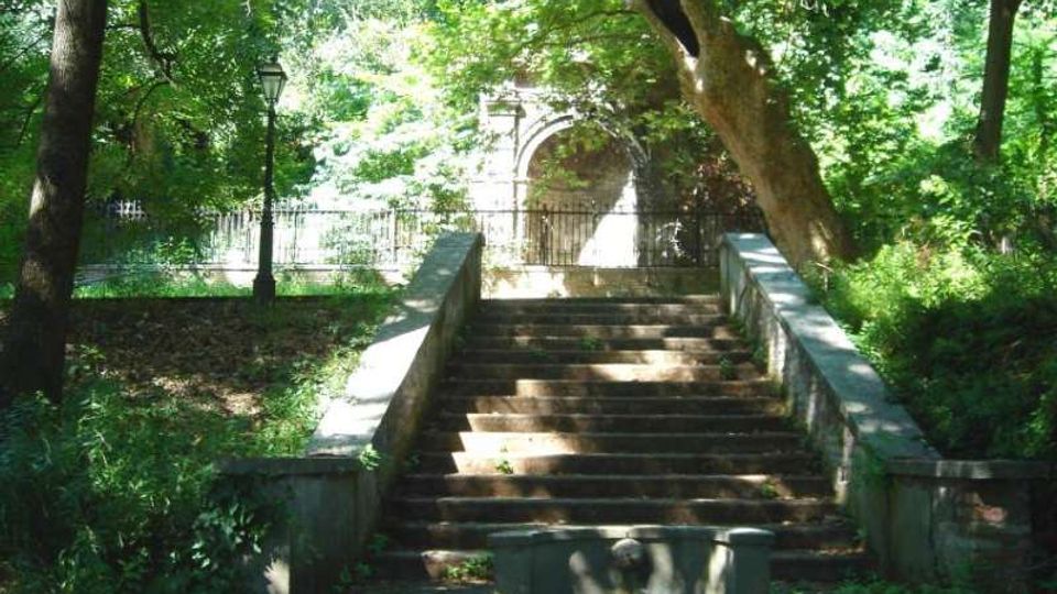 Scalinata in pietra in un parco alberato, con parapetti laterali e vegetazione fitta ai lati. In cima ai gradini si vede una recinzione metallica e, sullo sfondo, un arco in muratura parzialmente illuminato tra gli alberi.