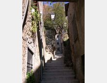 Scalinata in pietra in un vicolo stretto tra edifici in muratura, con pareti ruvide e finestre con grate. Un lampione in ferro sporge sopra le scale, con piante rampicanti e alberi che incorniciano la parte alta del passaggio.