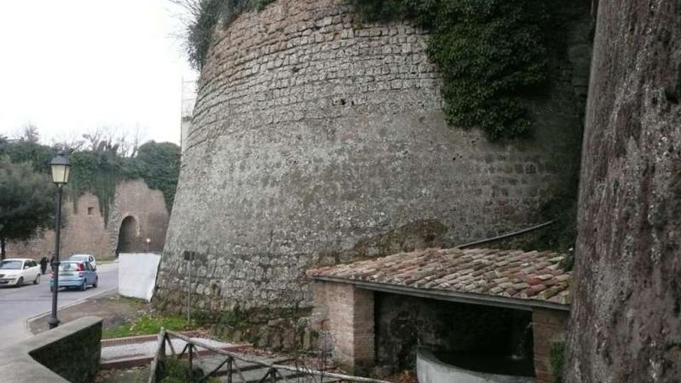 Muro di fortificazione in pietra con struttura semicilindrica e vegetazione sulla sommità, lungo una strada con lampione e auto parcheggiate. In primo piano si vede un piccolo edificio con tetto in coppi e una recinzione in legno su un percorso pedonale.
