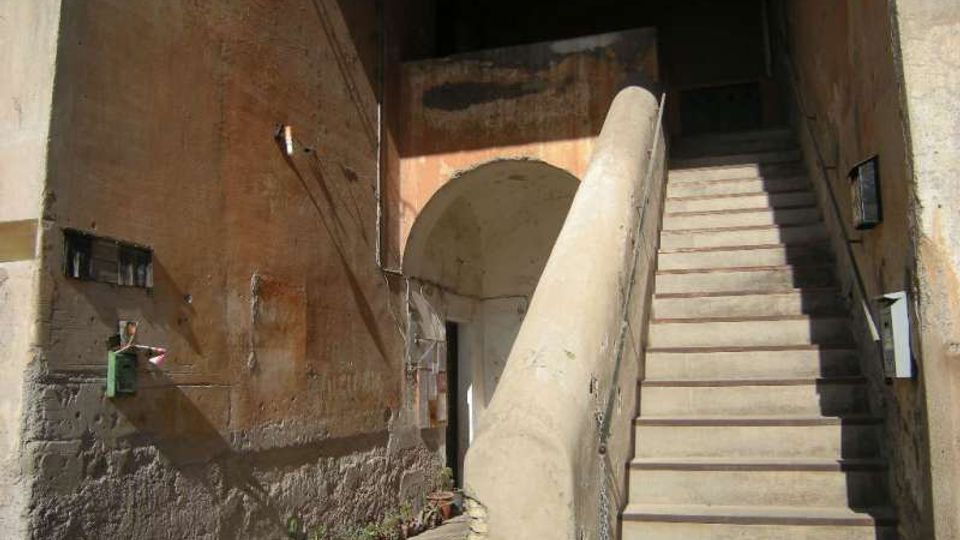 Cortile interno con pareti in intonaco consumato e un’ampia scala in pietra che sale a destra, affiancata da un alto parapetto. Sul fondo si vede un arco che conduce a un passaggio coperto e a un ingresso.