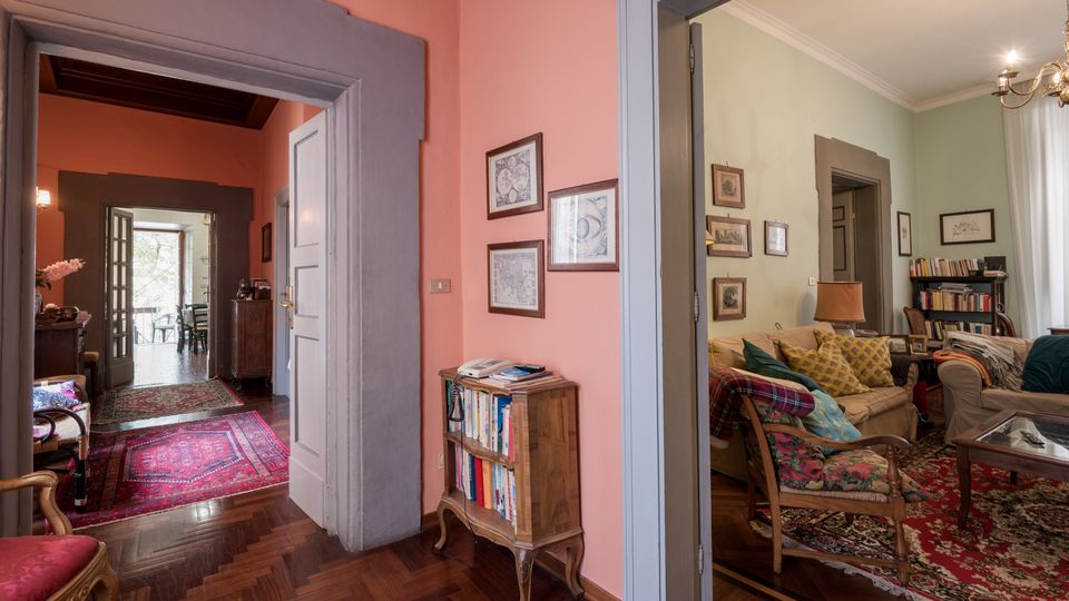 Interno di abitazione con corridoio dai muri rosa e pavimento in parquet, incorniciato da ampie porte con cornici grigie e tappeti persiani. A destra si apre un soggiorno con divani, poltrona, tavolino, libreria e lampada, illuminato da una finestra con tende chiare.