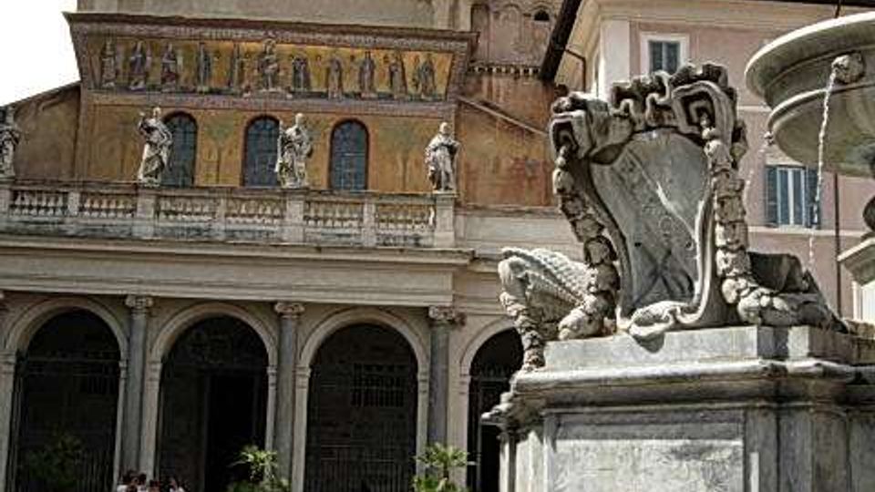Piazza con scalinata in primo piano e fontana monumentale in pietra decorata. Sullo sfondo si vede una chiesa con portico ad archi, facciata con mosaico e statue, e un campanile con orologio.