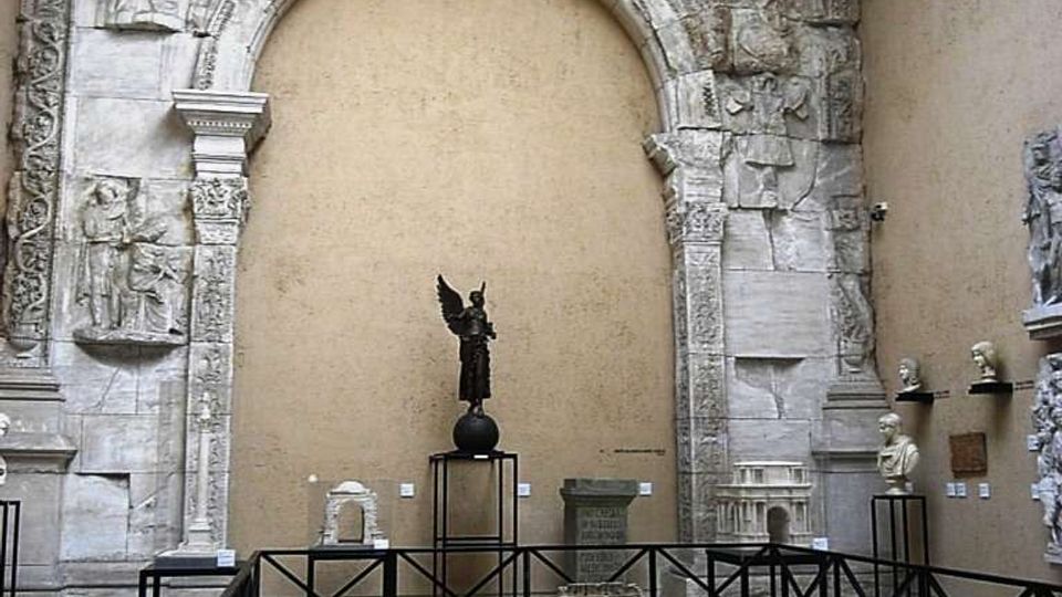 Sala museale con grande arco in pietra scolpita e iscrizioni, incassato in una parete color ocra e illuminato da un lucernario. Davanti all’arco sono esposti reperti e statue su piedistalli, con una struttura metallica che delimita una grande base circolare al centro.