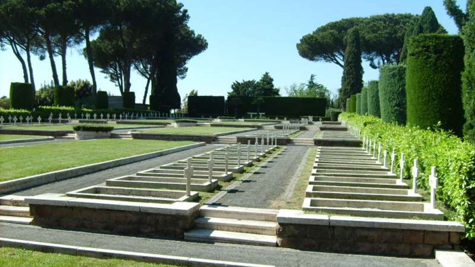 Cimitero all’aperto con file ordinate di piccole lapidi bianche disposte su terrazze in pietra e vialetti rettilinei. Sullo sfondo ci sono alte siepi potate e grandi pini a ombrello sotto un cielo sereno.
