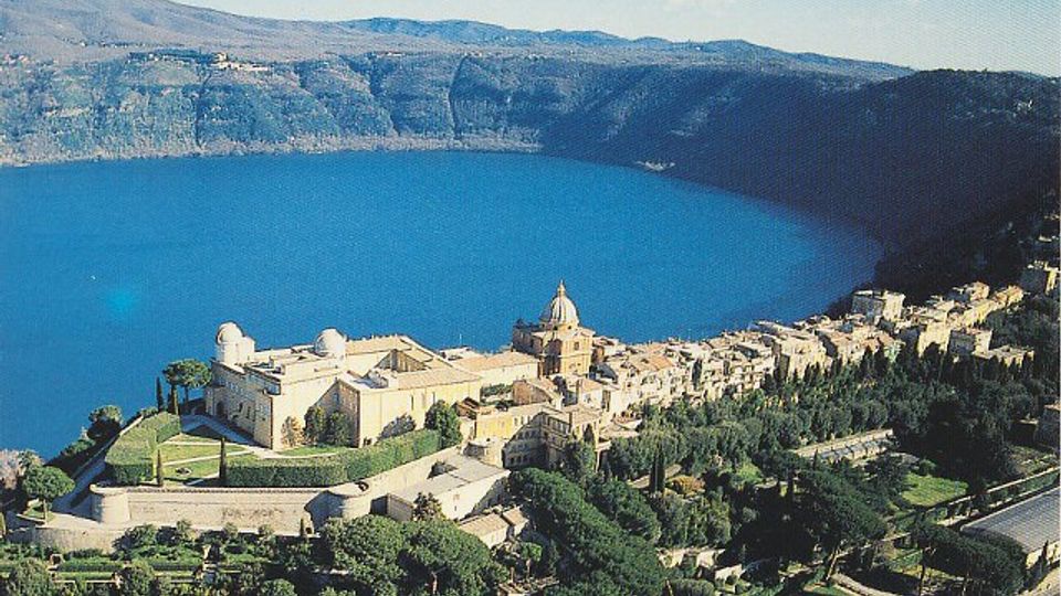 Veduta panoramica di un borgo affacciato su un ampio lago circondato da colline, con un grande complesso storico in primo piano caratterizzato da cupola e mura. Case in pietra e vegetazione terrazzata degradano verso la riva lungo una strada costiera.