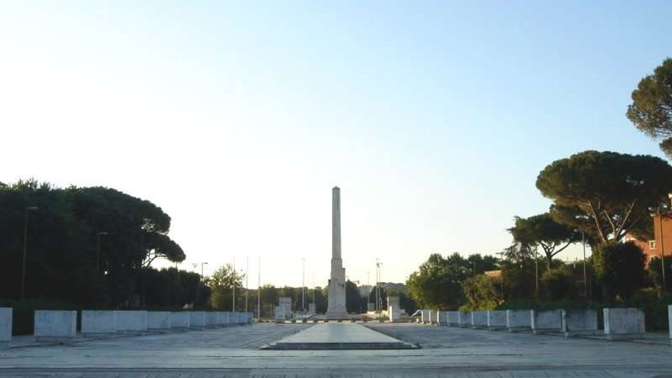 Ampia piazza monumentale pavimentata con decorazioni geometriche e figurative, delimitata da bassi parapetti e alberi ai lati. Al centro si erge un alto obelisco in pietra lungo un asse prospettico verso l’orizzonte.