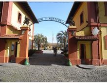 Ingresso monumentale a un complesso militare, con due edifici simmetrici giallo e rosso e un arco con la scritta “... di Montebello” tra due pilastri decorati. Davanti c’è una pavimentazione in ciottoli e oltre l’arco si vede un viale con palme e una bandiera al centro del cortile.