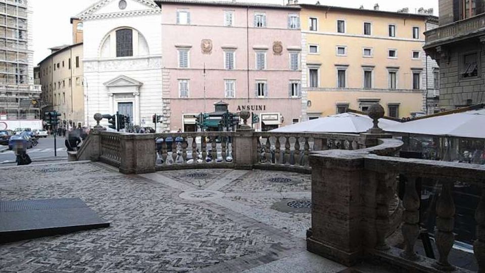 Veduta di una piazza urbana con pavimentazione in pietra e una terrazza rialzata con balaustra in muratura. Sullo sfondo si vedono una chiesa con facciata bianca e edifici storici color pastello, oltre a una strada con traffico e strutture coperte tipo dehors.
