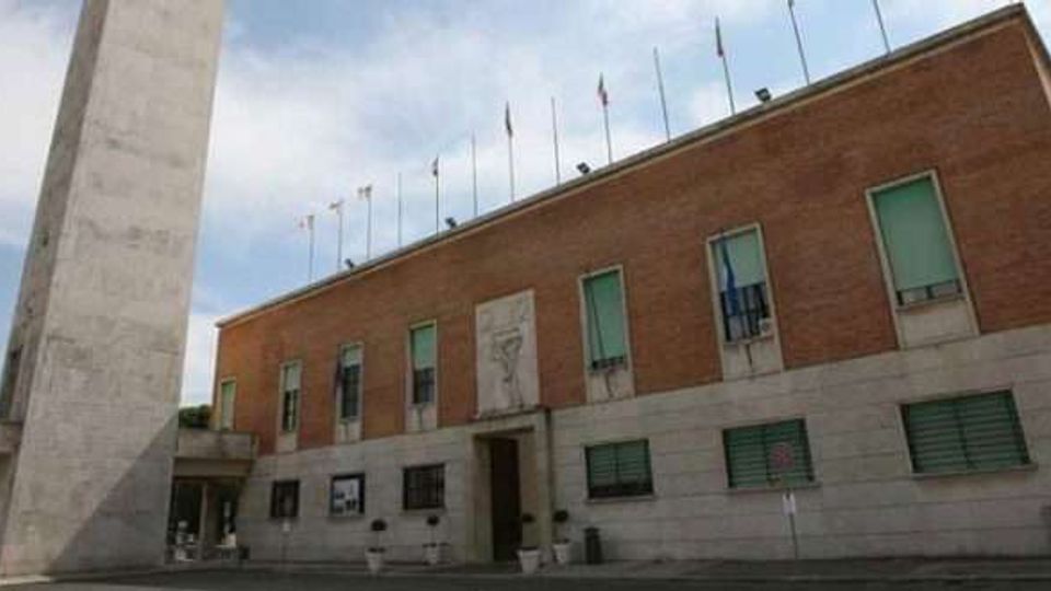 Facciata di un edificio istituzionale in mattoni rossi con basamento in pietra chiara, finestre rettangolari e ingresso centrale, con una fila di pennoni sul tetto. In primo piano si vede un alto obelisco in pietra e un’area pavimentata davanti all’edificio.