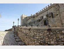 Castello medievale in pietra con mura merlate e torre, visto da una strada in salita lastricata e affiancata da un muretto in pietra e lampioni. Un grande arco d’ingresso si apre nella fortificazione sotto un cielo sereno.