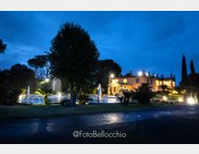 Veduta serale di una villa a due piani illuminata, circondata da un giardino con palme e alberi alti. In primo piano si vede una grande fontana con getti d’acqua e luci, accanto a un vialetto asfaltato bagnato.