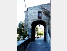 Passaggio ad arco sotto un edificio in pietra con facciata irregolare, finestra con balconcino e una lanterna sospesa al centro della volta. La strada acciottolata attraversa l’arco e conduce verso un’area con alberi e auto parcheggiate, con muretto laterale e cavi a vista sulla parete.