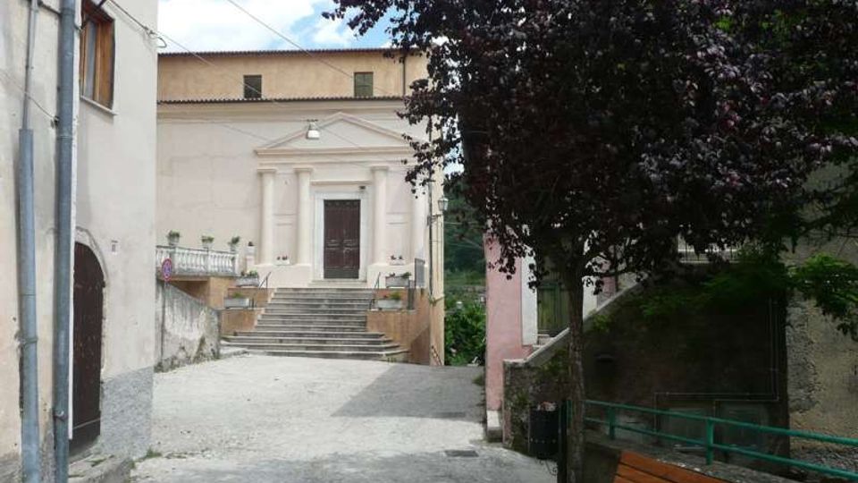 Stradina in salita di un borgo che conduce a un edificio con facciata classica, portale centrale e ampia scalinata in pietra. Ai lati ci sono case addossate, un grande albero che ombreggia il percorso, ringhiere e una panchina lungo il muro.