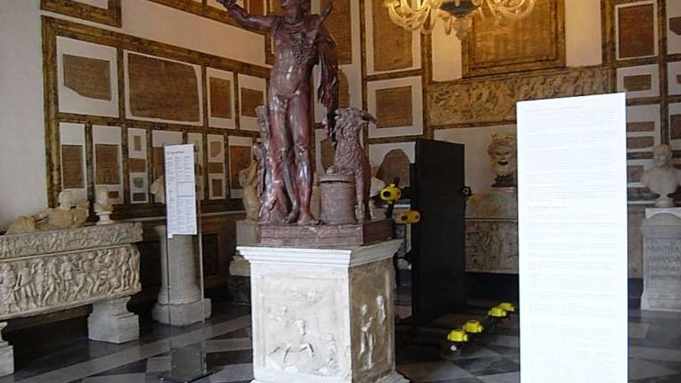 Sala museale con pavimento a scacchiera in marmo e soffitto decorato, illuminata da un grande lampadario. Al centro una statua su piedistallo, con pareti rivestite da lapidi e rilievi e pannelli informativi espositivi.