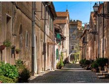 Stradina stretta acciottolata in un borgo storico, fiancheggiata da case in pietra con intonaco consumato, persiane e balconcini. Lungo i lati ci sono vasi di piante, lampioni a parete e, sullo sfondo, un arco e un edificio con tetto in tegole sotto un cielo sereno.