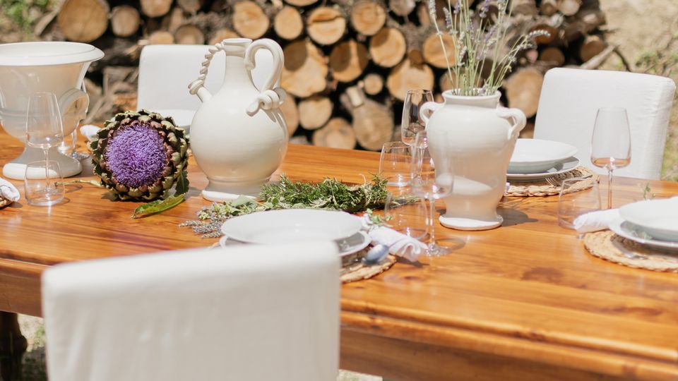Tavolo da pranzo in legno apparecchiato all’aperto con piatti bianchi, calici, brocche e vasi in ceramica con fiori, circondato da sedie con coperture bianche. Sullo sfondo si vede una catasta di tronchi tagliati contro una scarpata di terra.