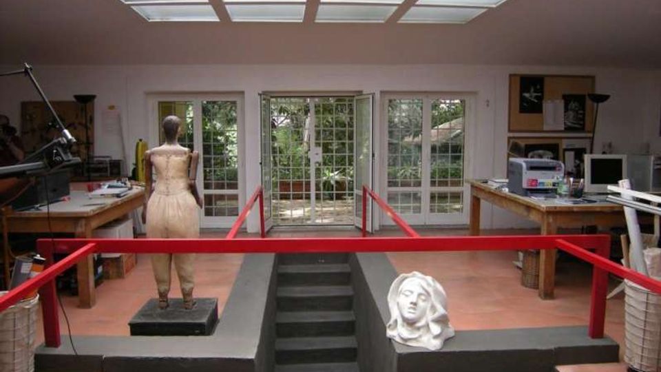 Interno di un laboratorio o studio artistico con lucernario a griglia sul soffitto, porte-finestre vetrate sul fondo e tavoli da lavoro ai lati. Al centro c’è una piattaforma rialzata con scala e parapetti rossi, con un manichino e una scultura di testa appoggiata.