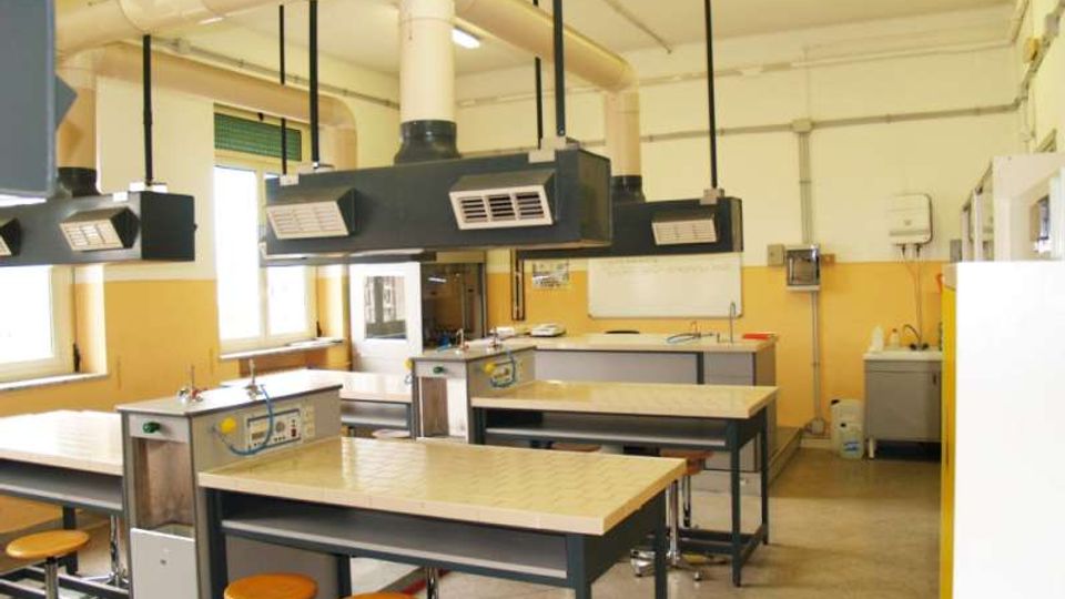 Aula laboratorio con grandi banchi da lavoro piastrellati e sgabelli, dotata di cappe aspiranti sospese e tubazioni di ventilazione a soffitto. Lungo le pareti sono presenti finestre, lavelli e attrezzature tecniche tipiche di un laboratorio scolastico o scientifico.