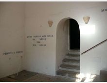 Interno di un edificio storico con pareti bianche e scritte murali; un arco conduce a una breve scalinata in pietra verso un corridoio buio. Sulla parete destra sono presenti una ringhiera e piccole applique, con luce naturale che entra da un’apertura fuori campo.