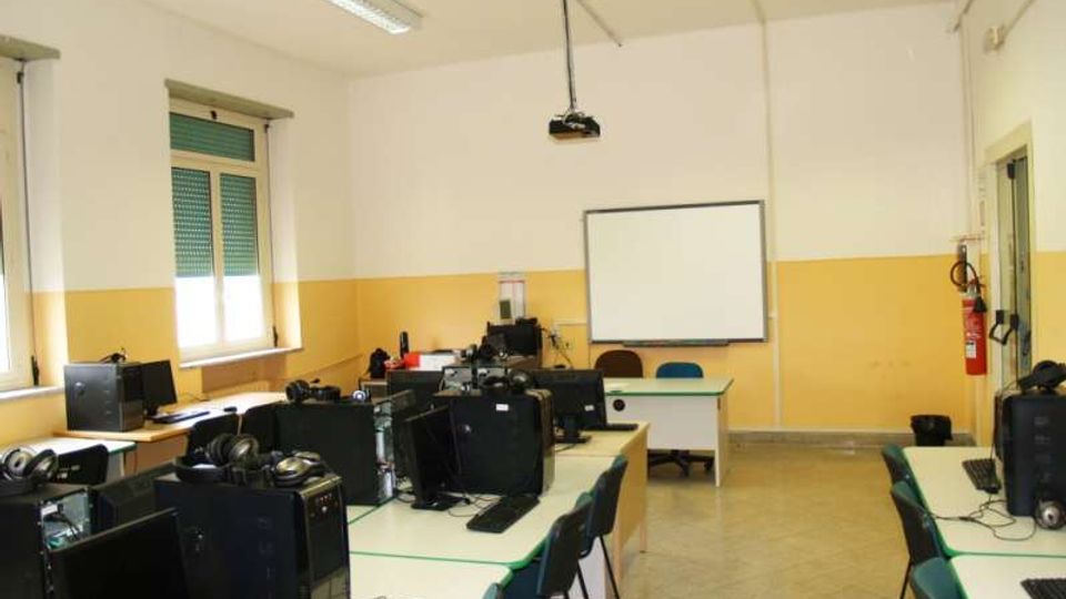 Aula informatica con banchi disposti a isole e postazioni con computer e monitor; sul fondo c’è una lavagna bianca e un proiettore a soffitto. Pareti bicolore bianco e giallo, finestre laterali con tapparelle e illuminazione a neon.