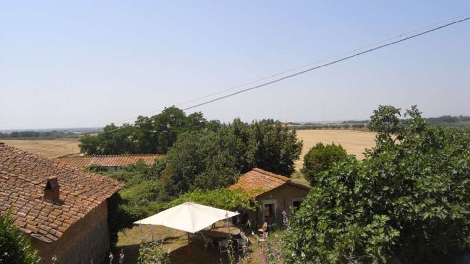 Vista di una casa di campagna con tetti in coppi e un cortile con gazebo bianco, circondata da alberi. Sullo sfondo si estendono campi coltivati e colline sotto un cielo limpido, con cavi elettrici che attraversano la parte alta dell’immagine.
