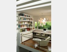 Interno di uno studio domestico moderno con pavimento in legno, libreria a parete e lunga scrivania sotto una grande finestra affacciata sul verde. Il soffitto ha lucernari a fasce e una lampada a sospensione con fogli appesi, con ringhiera metallica lungo il bordo del soppalco.