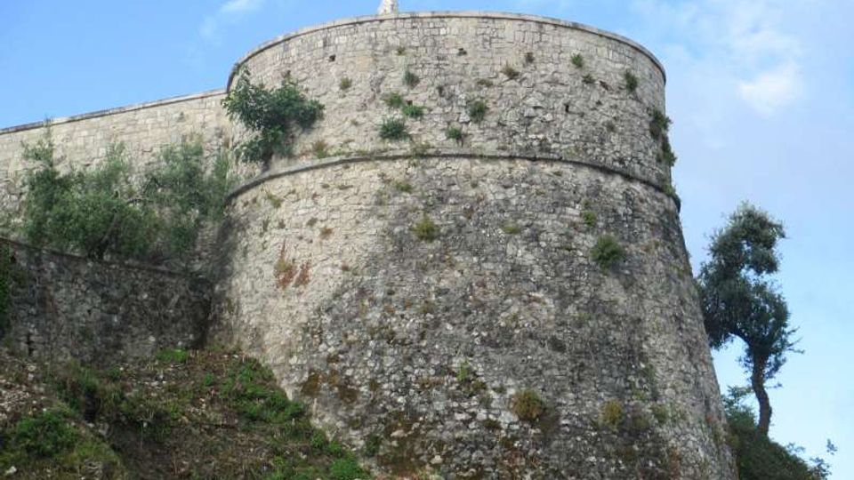 Torre circolare di una fortificazione in pietra con mura alte e spesse, vista dal basso su un pendio roccioso. Vegetazione spontanea cresce sulle pareti e intorno alla struttura, con cielo azzurro sullo sfondo.
