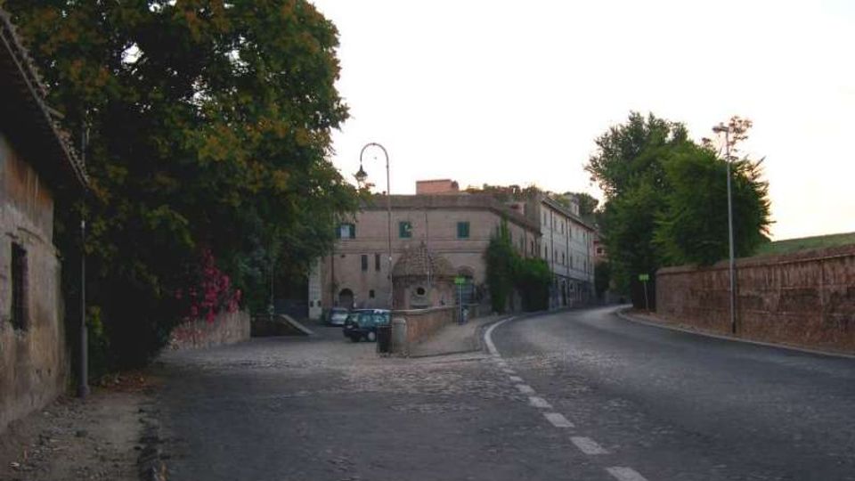 Strada asfaltata che curva a destra accanto a un marciapiede in pietra, con muri di mattoni e vegetazione ai lati. Sullo sfondo si vedono edifici in muratura chiara, un lampione ad arco e alcuni veicoli parcheggiati vicino a un piccolo accesso.