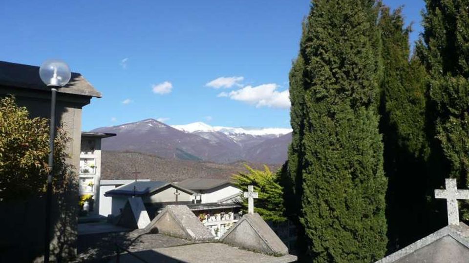 Cimitero su un pendio con tombe in pietra e croci, affiancato da alti cipressi e da un edificio laterale. Sullo sfondo si vede una valle con montagne innevate sotto un cielo sereno.