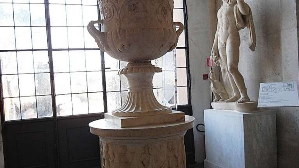 Sala museale con grande finestra a griglia e pavimento in marmo; al centro un grande cratere in pietra decorato su piedistallo, con una statua classica su basamento lungo la parete.